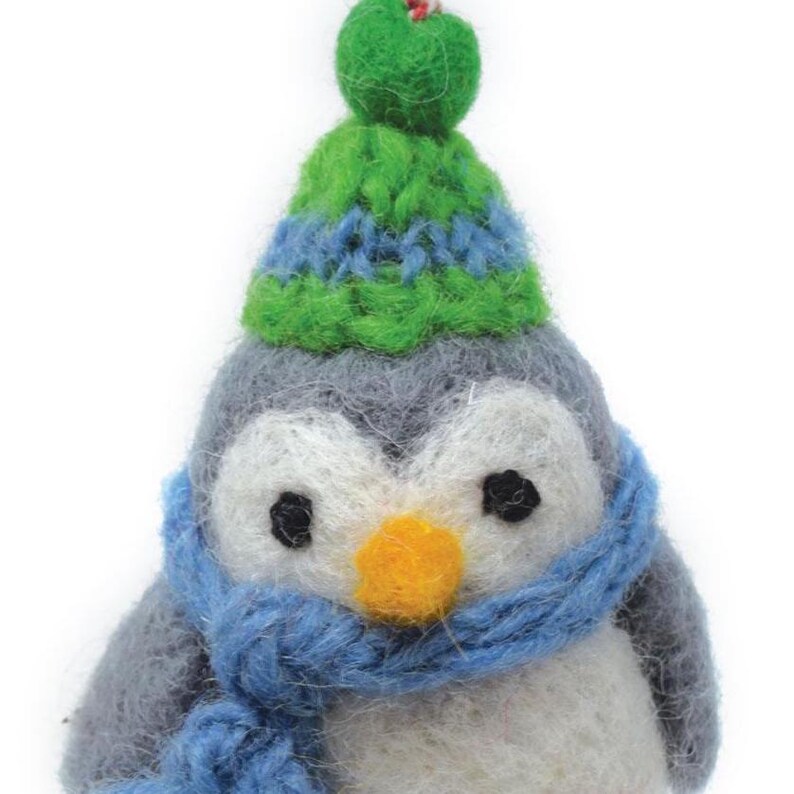 May include: A gray and white felt penguin wearing a green and blue knit hat and a blue scarf. The penguin has black eyes and an orange beak.