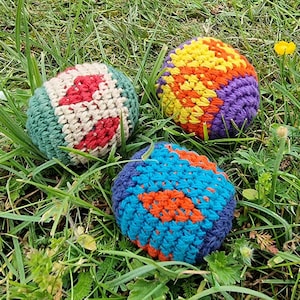 May include: Three colorful crocheted balls rest in green grass. One ball is primarily green, cream, and red. Another is yellow, orange, and purple. The third ball is blue and orange. These handmade balls are likely for play or decoration.