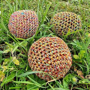 May include: Three colourful, crocheted spheres rest in green grass. The spheres are made of tightly woven yarn in a variety of colours, including red, yellow, blue, and green. The spheres appear to be handmade and have a textured surface.