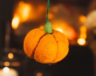 May include: A felt orange pumpkin ornament with a green stem hanging from a string. The pumpkin is in focus and the background is blurry with warm lights.