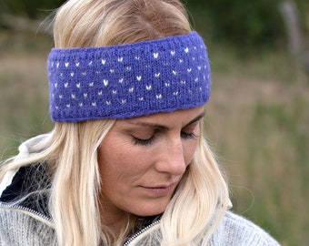 May include: A knitted blue headband with a white heart pattern. The headband is worn by a person with blonde hair and a gray sweater. The accessory is designed to keep the ears and forehead warm.