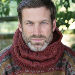 May include: A thick, ribbed, rust-colored knit cowl neck scarf worn over a multi-colored, patterned wool sweater. The scarf features a chunky, textured knit construction with visible vertical ribbing, providing a warm, layered appearance.