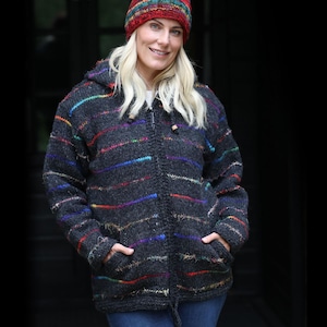 May include: A dark gray hooded jacket with colorful horizontal stripes. The jacket has a zipper closure, two front pockets, and a drawstring at the hood. The person is also wearing a multicolored knit hat.