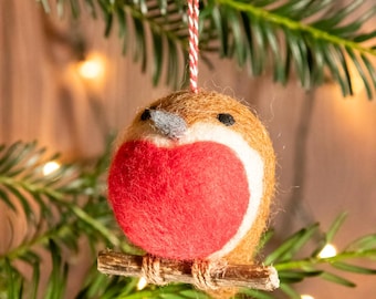 May include: A felted Christmas ornament shaped like a robin bird. The bird has a red breast, brown body, and a small wooden perch. It hangs from a red and white string, set against a backdrop of evergreen branches and soft lights.