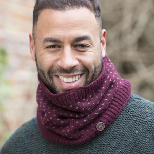 May include: A burgundy knit cowl with a button closure and a dotted pattern. The cowl is worn over a green sweater. The image is a close-up shot of the cowl and the person wearing it.