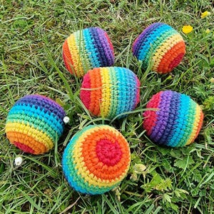 May include: Six colorful, crocheted balls in a rainbow of hues, including red, orange, yellow, green, blue, and purple, are scattered on a bed of green grass. The balls are round and appear to be handmade, showcasing a vibrant, playful design.
