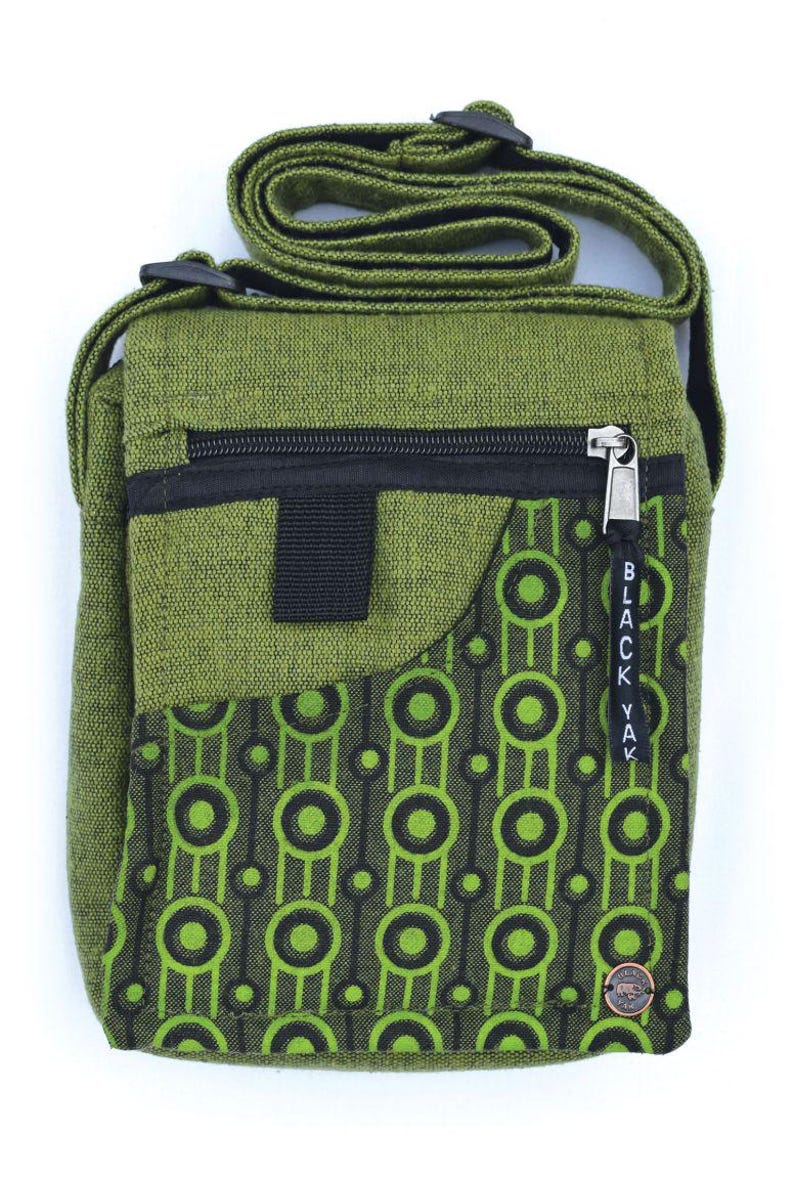May include: A green canvas crossbody bag with a black zipper and a black and green geometric pattern on the front flap. The bag has a black strap and a small black pocket on the front. The bag has a black "BLACK YAK" tag hanging from the zipper.