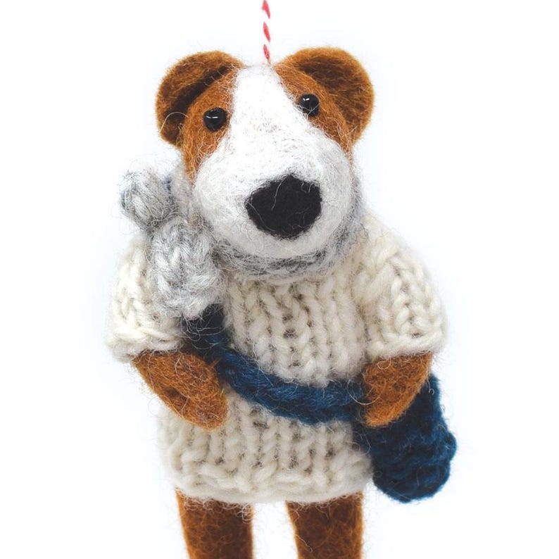 Puede incluir: Un adorno de perro de fieltro que lleva un su&eacute;ter de punto blanco con una bufanda azul y una bolsa azul. El perro tiene pelo marr&oacute;n y blanco y una nariz negra.