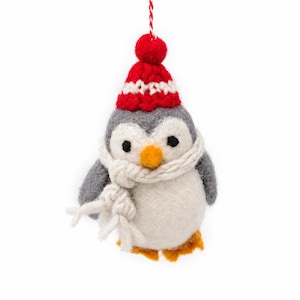 May include: A felted wool penguin ornament featuring a gray and white body, a bright orange beak, and orange feet. The penguin wears a red and white knit hat with a pom-pom on top and a thick, cream-colored yarn scarf wrapped around its neck.