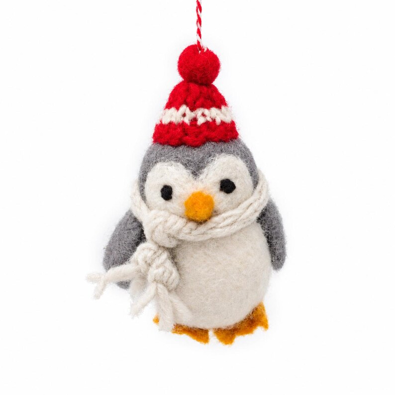 May include: A felted wool penguin ornament featuring a gray and white body, a bright orange beak, and orange feet. The penguin wears a red and white knit hat with a pom-pom on top and a thick, cream-colored yarn scarf wrapped around its neck.