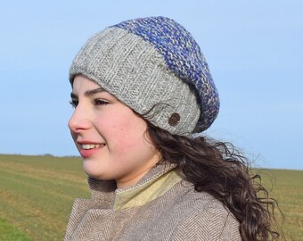 May include: A gray and blue knit beanie with a button on the side. The beanie is worn by a person with curly brown hair.