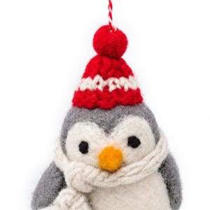 May include: A felted wool penguin ornament featuring a gray and white body, a bright orange beak, and black eyes. The penguin wears a red and white knit hat with a pom-pom on top and a thick, cream-colored knit scarf wrapped around its neck.
