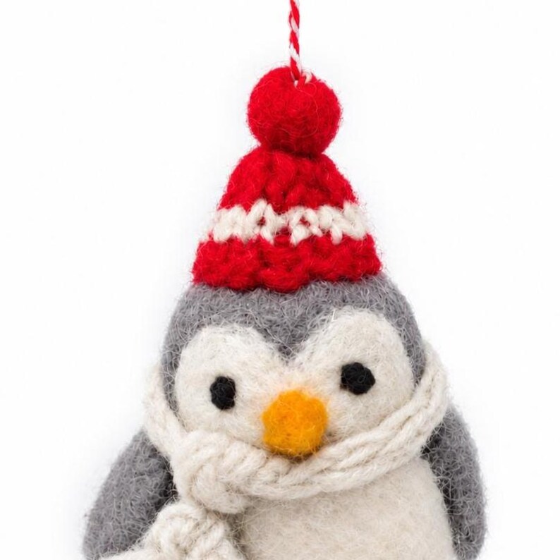 May include: A felted wool penguin ornament featuring a gray and white body, a bright orange beak, and black eyes. The penguin wears a red and white knit hat with a pom-pom on top and a thick, cream-colored knit scarf wrapped around its neck.