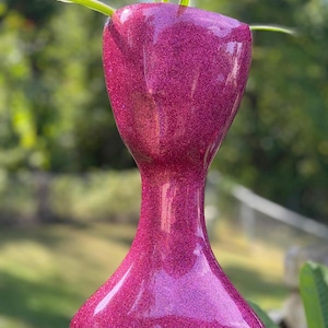 May include: A tall, hourglass-shaped planter with a vibrant pink glitter finish. The planter holds green plants with long, slender leaves. The planter is set outdoors against a blurred green background.