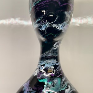 May include: A black decorative vase with a unique, swirling paint design. The vase features a blend of colors, including white, teal, and purple, creating an abstract, marbled effect. The vase has a wide base, a slender neck, and a flared top.