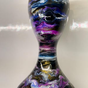 May include: A black hourglass-shaped decorative vase with a swirling pattern of purple, blue, white, and gold. The vase has a wide base, a narrow middle, and a flared top, showcasing an abstract design.