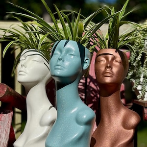 May include: Three decorative head planters in white, teal, and bronze, each holding a vibrant green plant. The planters are shaped like stylized human heads with closed eyes and are placed outdoors.