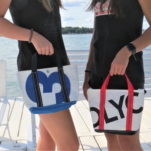 May include: Two white canvas tote bags with contrasting colored handles and lettering. The bag on the left has a blue handle and blue lettering that says "M". The bag on the right has a red handle and red lettering that says "OLY".