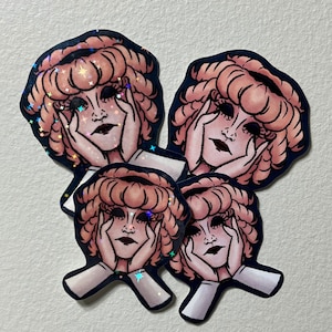 May include: A collection of stickers featuring a stylized illustration of a person with peach-coloured curly hair, dark eyes, and hands supporting their face. The stickers have a dark blue border and a holographic, glittery finish.