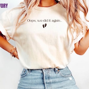 May include: An ivory short-sleeved t-shirt featuring the black printed text "Oops, we did it again..." centred on the chest, positioned above a small graphic of two dark baby footprints. The shirt has a crew neckline and a relaxed fit.