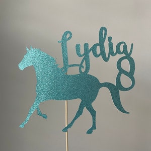 May include: A teal glitter horse cake topper with the name "Lydia" and the number "8". The horse is in a running pose and is attached to a wooden stick.