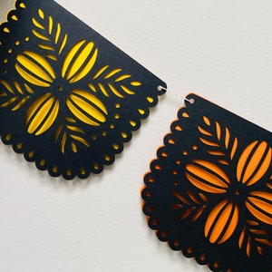 Puede incluir: Una pancarta decorativa con dise&ntilde;os recortados. El papel negro tiene un borde festoneado y intrincados patrones florales con respaldo amarillo y naranja. La pancarta est&aacute; unida con una cuerda blanca.