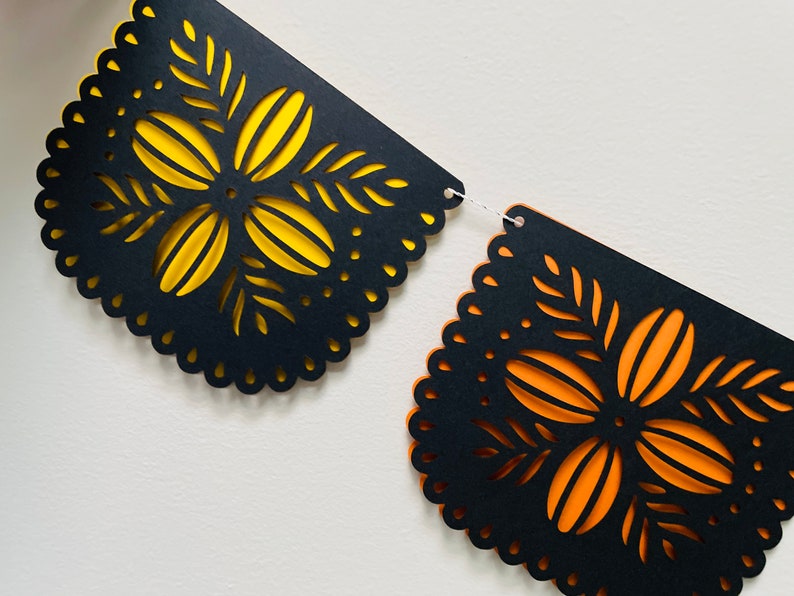 Puede incluir: Una pancarta decorativa con dise&ntilde;os recortados. El papel negro tiene un borde festoneado y intrincados patrones florales con respaldo amarillo y naranja. La pancarta est&aacute; unida con una cuerda blanca.