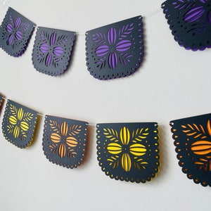 Puede incluir: Una guirnalda decorativa con bordes festoneados y recortes florales. La guirnalda presenta secciones alternas de p&uacute;rpura y amarillo superpuestas sobre papel negro. La guirnalda est&aacute; ensartada en una cuerda blanca, adecuada para ocasiones festivas.