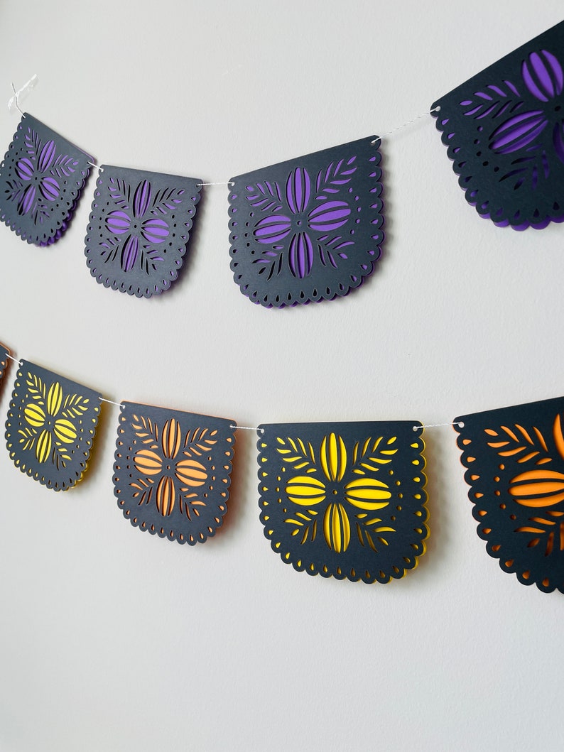 Puede incluir: Una guirnalda decorativa con bordes festoneados y recortes florales. La guirnalda presenta secciones alternas de p&uacute;rpura y amarillo superpuestas sobre papel negro. La guirnalda est&aacute; ensartada en una cuerda blanca, adecuada para ocasiones festivas.
