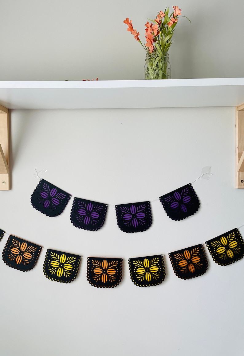 Puede incluir: Una guirnalda decorativa con banderines de papel negro cortados con l&aacute;ser. Cada bander&iacute;n presenta un dise&ntilde;o floral en tonos morado, naranja y amarillo. La guirnalda est&aacute; ensartada en una cuerda blanca, ideal para fiestas.