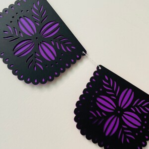 Puede incluir: Una guirnalda decorativa con dise&ntilde;os recortados en negro y un respaldo morado. La guirnalda tiene un borde festoneado y patrones florales. La guirnalda est&aacute; ensartada en una cuerda blanca, adecuada para ocasiones festivas.