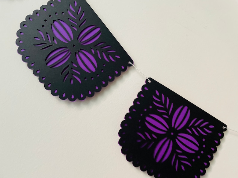 Puede incluir: Una guirnalda decorativa con dise&ntilde;os recortados en negro y un respaldo morado. La guirnalda tiene un borde festoneado y patrones florales. La guirnalda est&aacute; ensartada en una cuerda blanca, adecuada para ocasiones festivas.