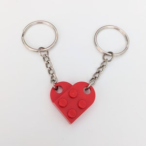 Puede incluir: Un llavero de corazón rojo Lego con dos llaveros plateados conectados por una cadena plateada.