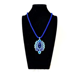 Può includere: Una collana con pendente blu su un espositore nero. Il pendente presenta una grande gemma ovale blu scuro, circondata da intricati dettagli blu chiaro e argento. La collana ha un cordino blu abbinato.