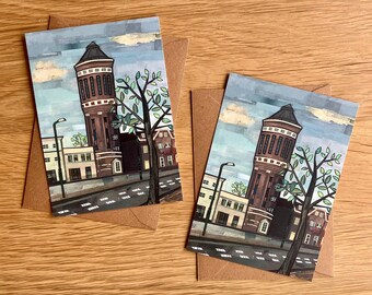 May include: Two illustrated greeting cards featuring a red brick water tower with a green tree in front of it. The cards are on a brown wooden surface.