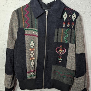May include: A patchwork jacket with a navy blue collar and a silver zipper. The jacket features various fabric panels in gray, black, and brown, adorned with colorful embroidered patterns and designs. The jacket has a ribbed hem and cuffs.