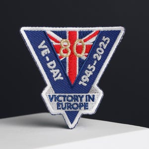 Puede incluir: Un parche bordado en azul, rojo y blanco con un diseño de Union Jack. El parche tiene forma de triángulo y tiene el texto "VE-DAY 1945-2025" y "VICTORY IN EUROPE" en él. El número "80" está en el centro del parche.