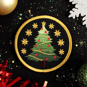 Pode incluir: Um remendo bordado de Natal com uma árvore de Natal dourada com detalhes verdes e vermelhos sobre um fundo preto. O remendo é rodeado por glitter dourado e um floco de neve branco.