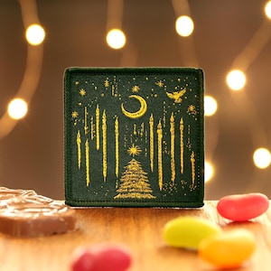 May include: A green fabric patch with gold embroidery depicting a crescent moon, stars, a Christmas tree, and a flock of birds. The patch is a festive design for a holiday project.