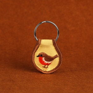 Könnte beinhalten: Ein gelber Stoffschlüsselanhänger mit einem braunen gestickten Rotkehlchen-Design. Der Schlüsselanhänger hat einen silbernen Metallring.