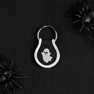 May include: A white ghost keychain with a black background and a white border. The keychain is attached to a silver ring.