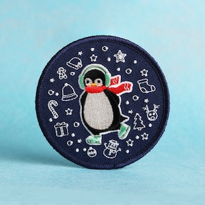 Puede incluir: Un parche circular azul con detalles bordados en blanco de un pingüino que lleva una bufanda roja y auriculares, rodeado de copos de nieve, estrellas y otros símbolos navideños de invierno.