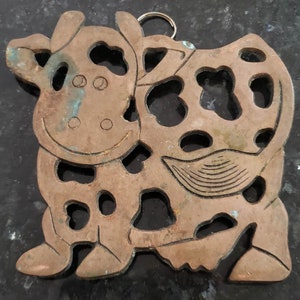 May include: A cutout of a cow made of copper with a loop at the top for hanging. The cow is brown with a green patina and has a detailed design.