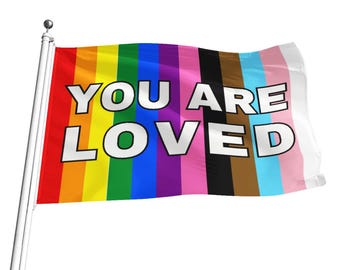 Puede incluir: Una bandera del orgullo arcoíris con las palabras "YOU ARE LOVED" en letras blancas. La bandera está hecha de una tela suave y fluida y ondea en un asta de bandera.