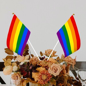 May include: Two rainbow pride flags on white sticks, placed in a bouquet of brown and orange flowers. The flags are made of fabric and have six stripes of red, orange, yellow, green, blue, and purple.