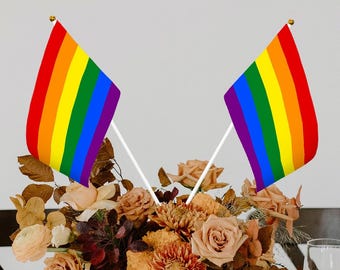 Puede incluir: Dos banderas arcoíris del orgullo en palos blancos, colocadas en un ramo de flores marrones y naranjas. Las banderas están hechas de tela y tienen seis franjas de rojo, naranja, amarillo, verde, azul y morado.