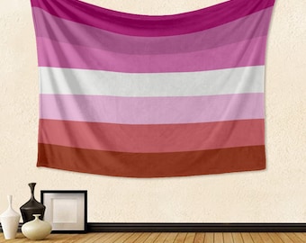 Puede incluir: Una bandera del orgullo lésbico colgada en una pared blanca. La bandera tiene seis franjas horizontales en los colores rosa, rosa claro, blanco, morado claro, rojo y marrón.