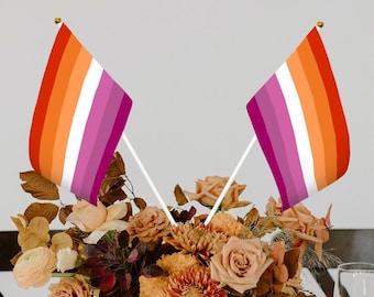 Puede incluir: Dos pequeñas banderas del orgullo lésbico con rayas naranjas, rosas y blancas se exhiben en un arreglo floral. Las banderas están sobre palos blancos con remates dorados. Las flores son en tonos naranja, rosa y marrón.