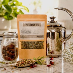 May include: A tan pouch of organic pregnancy tea by Wild Roots Tea Co. with a clear window showing dried herbs, positioned next to a glass mason jar filled with dried fruit, a silver metal tea kettle, and a metal spoon containing loose tea leaves.