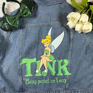 Op de afbeelding: Een blauwe denimjas met een groene Tinkerbell-illustratie en de tekst "TINK Being perfect isn't easy".